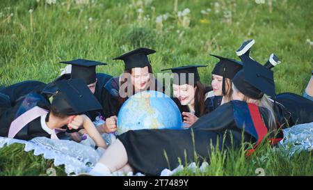 Diplômés en robes noires regardant un globe géorgraphique allongé sur l'herbe. Banque D'Images