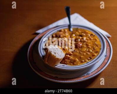 FABADA ESPAGNOLE TYPIQUE DANS UNE ASSIETTE, AVEC UN MORCEAU DE PAIN ET UNE CUILLÈRE DESSUS. TOUT EST SUR UNE TABLE EN BOIS AVEC UNE SERVIETTE Banque D'Images