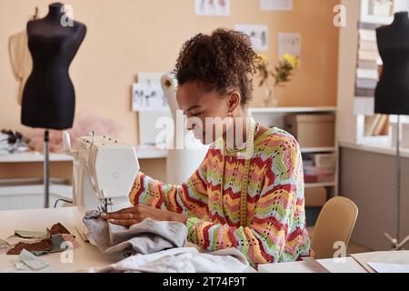 Jolie jeune créatrice de mode féminine assise par la machine à coudre électrique et créant de nouveaux articles pour la collection saisonnière ou upcycling vieux vêtements Banque D'Images