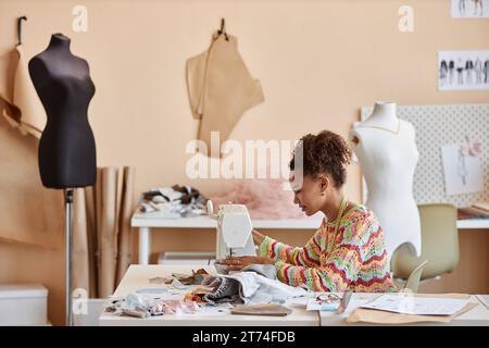 Long plan de la jeune femme afro-américaine artisan upcycling vieux vêtements dans l'atelier tout en étant assis par la machine à coudre électrique devant la caméra Banque D'Images