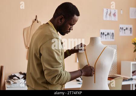 Vue latérale du jeune homme noir mettant du ruban adhésif sur la poitrine du mannequin tout en faisant contour du nouveau blazer de la collection saisonnière Banque D'Images