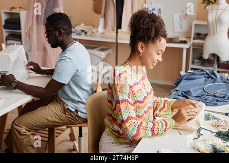 Vue latérale de deux collègues assis près des tables dans l'atelier et travaillant sur le recyclage de vieux vêtements pendant que l'un d'eux fait de la broderie Banque D'Images