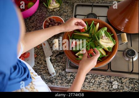 Vue d'en haut d'une femme musulmane du Moyen-Orient en hijab bleu, cuisinant délicieux repas végétarien dans un pot d'argile tajine, debout près du comptoir de cuisine à h. Banque D'Images