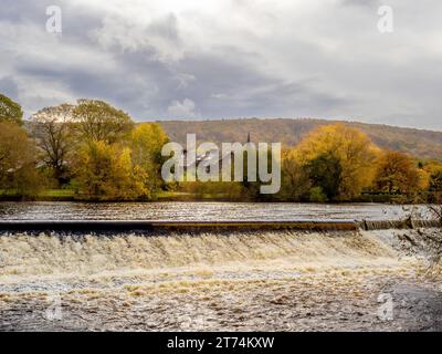 Le déversoir sur la rivière Wharfe à Otley, avec le paysage automnal du parc forestier Otley Chevin s'élevant au loin. Banque D'Images