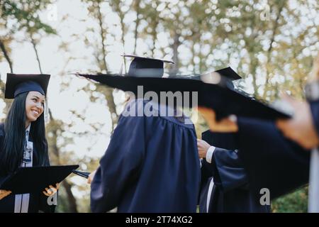 Divers étudiants universitaires vêtus de casquettes et de robes célèbrent l'obtention de leur diplôme par une journée ensoleillée. Ils marchent, s’amusent et se félicitent dans un parc. Banque D'Images