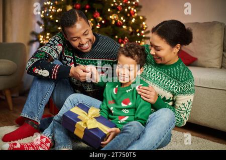 Joyeux parents afro-américains regardant avec amour leur fils pendant qu'il tient un cadeau de Noël Banque D'Images