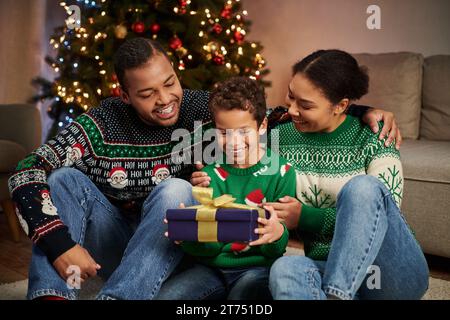 Joyeuse famille afro-américaine dans des pulls confortables regardant joyeusement le fils tenant le présent, Noël Banque D'Images