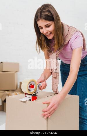 Jeune femme souriante tenant la machine à emballer scellant des boîtes en carton avec du ruban adhésif Banque D'Images