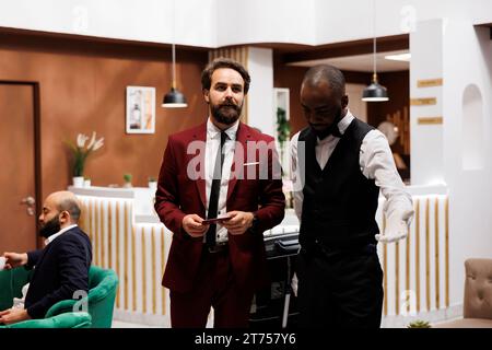 Assistant conduisant le client de l'hôtel à sa chambre, aide à transporter les bagages dans le hall après l'enregistrement. Bellhop afro-américain assurant un service de luxe et une assistance pour homme d'affaires formel. Banque D'Images