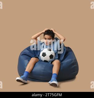 Petit garçon choqué avec un ballon de football assis dans une chaise de pouf sur fond beige Banque D'Images