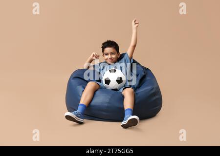 Heureux petit garçon avec ballon de football assis dans une chaise pouf sur fond beige Banque D'Images