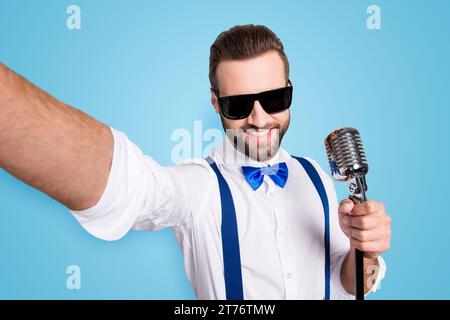 Autoportrait d'un chanteur élégant et élégant portant des bretelles de chemise en noeud papillon filant selfie sur l'appareil photo avant tenant le micro isolé sur fond gris Banque D'Images