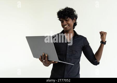 Heureux excité employé d'homme d'affaires indien tenant l'ordinateur portable debout isolé sur fond gris célébrant la victoire, de bons résultats en ligne, affaires Banque D'Images