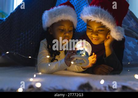 Garçon et fille afro-américains couchés et regardant la boule de neige illuminée de noël à la maison Banque D'Images