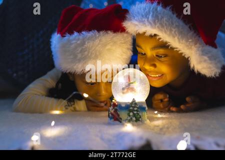 Garçon et fille afro-américains couchés et regardant la boule de neige illuminée de noël à la maison Banque D'Images