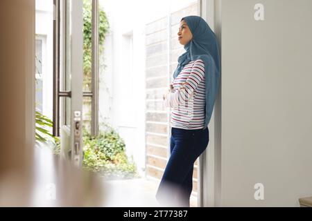 Femme biracial réfléchie en hijab bleu debout à la maison ensoleillée Banque D'Images