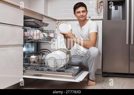 bel homme positif sort la vaisselle propre du lave-vaisselle dans la cuisine moderne blanche Banque D'Images