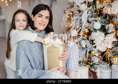 Mère et fille adorées partageant un moment tendre, tenant un cadeau avec un sapin de Noël festif en toile de fond Banque D'Images