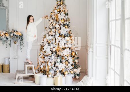 Une jeune fille dans un chandail blanc atteint joyeusement pour décorer un sapin de Noël somptueux dans une pièce lumineuse et élégante Banque D'Images