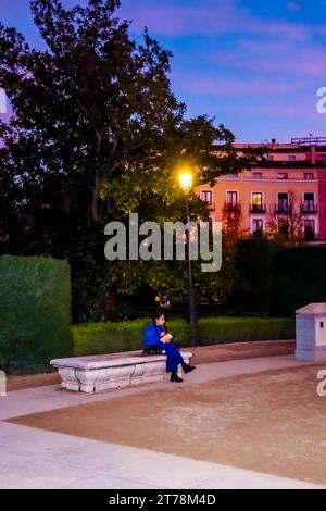 Femme assise sur un banc dans la soirée lisant par la lumière de la rue sur la Plaza de Oriente entourée d'arbres, Madrid Espagne. Banque D'Images