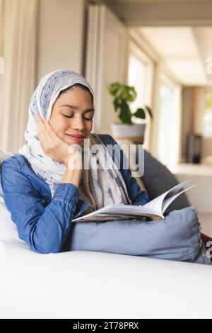 Heureuse femme biracial dans le livre de lecture de hijab dans le salon Banque D'Images