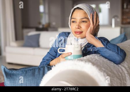 Femme biraciale dans le hijab buvant du café dans le salon Banque D'Images