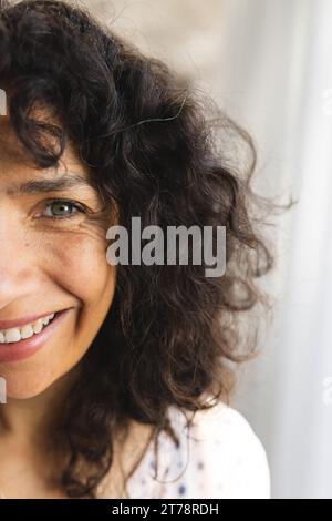 Demi portrait de femme mature caucasienne heureuse avec de longs cheveux noirs bouclés, souriant au soleil à la maison Banque D'Images