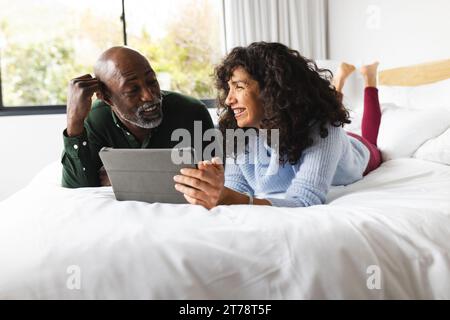 Heureux couple mûr diversifié couché sur le lit en utilisant tablette dans la chambre ensoleillée Banque D'Images