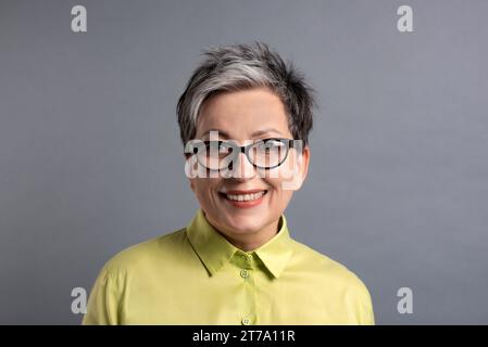 souriant femme âgée des années 50 dans des lunettes élégantes avec de courts cheveux gris regardant la caméra sur fond gris Banque D'Images