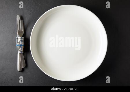 Assiette en céramique blanche vide avec couverts enveloppés dans un billet de cent dollars sur une table en pierre noire. Vue de dessus avec espace de copie Banque D'Images