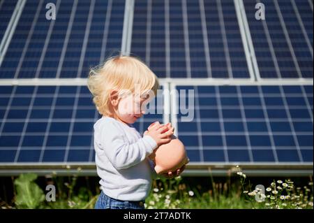 Petit enfant économisant de l'argent dans la tirelire sur fond de panneaux solaires. Enfant souriant heureux qu'il ait de l'argent dans sa caisse. Petit garçon apprenant à économiser de l'argent pour l'avenir. Banque D'Images