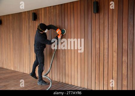 Travailleur de pont dans le masque ponçant le parement de bois dur avec ponceuse orbitale, terrasse en bois et rénovation de revêtement Banque D'Images