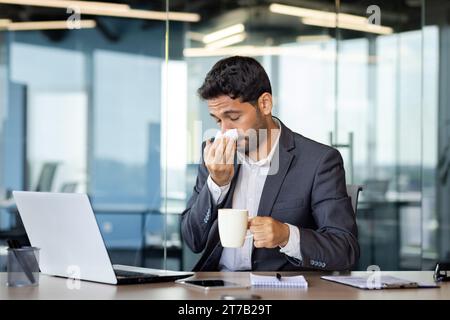 Jeune homme d'affaires hindou éternue et a un nez qui coule tout en travaillant dans le bureau à l'ordinateur, jeune homme a un rhume, boit des médicaments, se sent mal. Banque D'Images