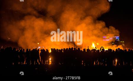 Un grand groupe de personnes s'est réuni autour d'un feu de joie sur une scène éclairée par les lumières brillantes des bâtiments en arrière-plan Banque D'Images