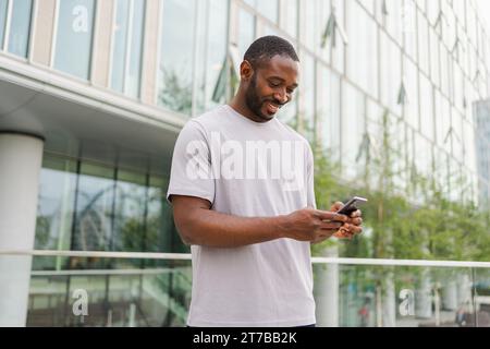 Homme afro-américain tenant l'écran tactile de smartphone tapant la page de défilement sur la rue urbaine dans la ville. Guy avec téléphone portable surfant sur Internet en utilisant des applications de médias sociaux magasinant en ligne. Addiction au téléphone portable Banque D'Images