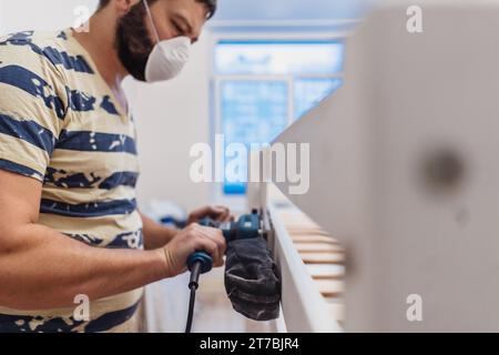 l'homme frotte le lit d'enfant en bois avec une ponceuse électrique ou un broyeur en enlevant la vieille peinture Banque D'Images