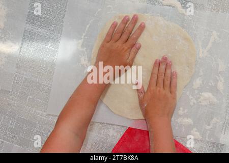 Gros plan : mains des enfants roulant la pâte en une couche. Il y a beaucoup de farine sur la table blanche. Banque D'Images