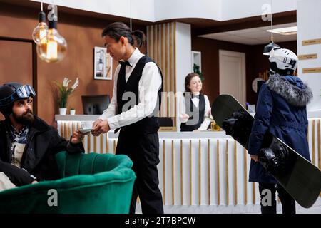 Arrivée d'une femme touriste transportant du snowboard dans le hall de l'hôtel pour l'enregistrement avec le personnel. Jeune serveur servant du café à l'homme sur le canapé. Ambiance de vacances d'hiver avec équipement de ski. La station de ski assure un séjour heureux. Banque D'Images