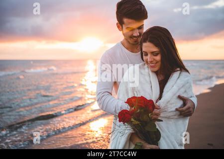 Couple souriant marchant plage avec bouquet roses coucher de soleil Banque D'Images