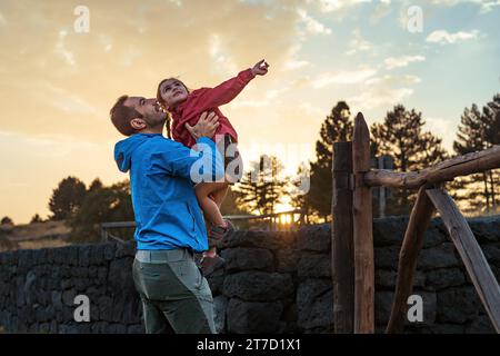 Père soulevant de manière ludique sa fille alors qu'ils profitent d'un coucher de soleil ensemble, capturant un joyeux moment de liens familiaux. Banque D'Images