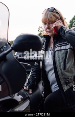 Portrait vertical de motard blonde mature qui parle sur le téléphone portable tout en étant assis sur sa moto. Communication dans les voyages. Hobby - moto t Banque D'Images