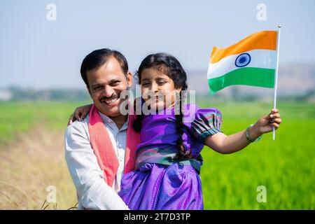 Joyeux père de village indien portant fille avec drapeau indien en marchant près de terres agricoles vertes pendant le jour de l'indépendance - concept de démocratie Banque D'Images