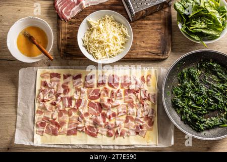 Vue de dessus du processus de préparation du mini gâteau à pizza. Bacon empilé sur pâte feuilletée, à côté des épinards, du fromage râpé et de l'œuf - vue de dessus. Recette traitée Banque D'Images