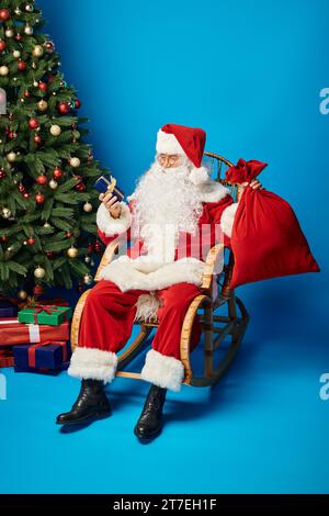 Joyeux Père Noël assis dans une chaise à bascule avec cadeau et sac à dos près de l'arbre de Noël sur bleu Banque D'Images