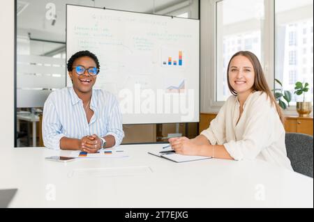 Ambiance de bureau conviviale. Deux collègues femmes riant en regardant la caméra, ayant une bonne communication au sein de l'équipe. Divers collègues dans la salle de réunion Banque D'Images