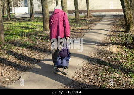 La femme porte un chariot. Le retraité traverse le parc. Un vieil homme va au magasin. Femme sur la piste en été. Banque D'Images