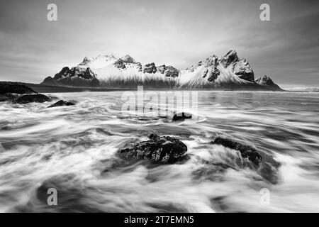 Image en noir et blanc d'une formation de montagne enneigée reflétée dans l'eau d'une plage de sable noir de lave, dans les vagues de premier plan lavées autour de b Banque D'Images