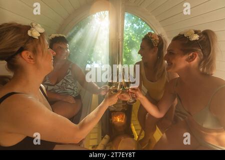 Bachelorette fête dans un sauna finlandais avec des femmes en maillots de bain et accessoires de cheveux floraux grillage du vin Banque D'Images