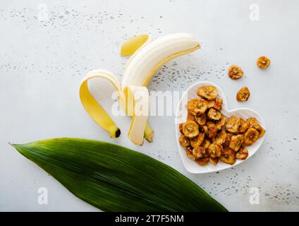 Tranches de banane séchées dans une assiette en forme de coeur avec banane crue et feuille verte.vue de dessus. Banque D'Images