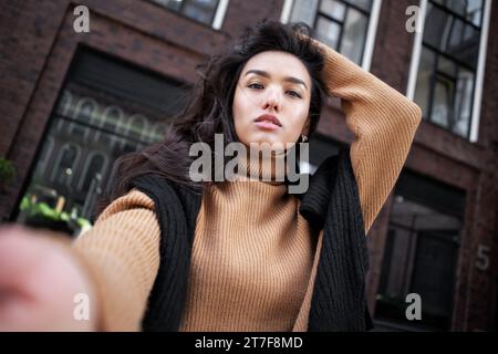 Femme attrayante avec les cheveux foncés prend le portrait Selfie, regardant la caméra à l'extérieur sur le fond du bâtiment. Beau modèle de mode féminin sur la rue de Banque D'Images